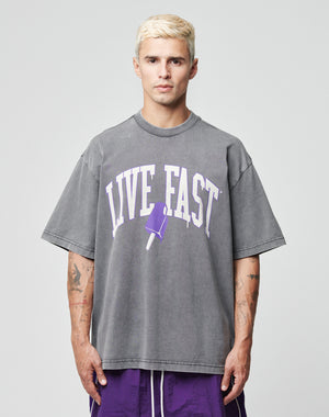 A person with short blond hair wears the LIVE FAST DIE YOUNG Live Fast Ice Tee, an oversized grey 100% Baumwolle shirt with a purple popsicle graphic and LIVE FAST print, paired with purple shorts against a plain light gray background.