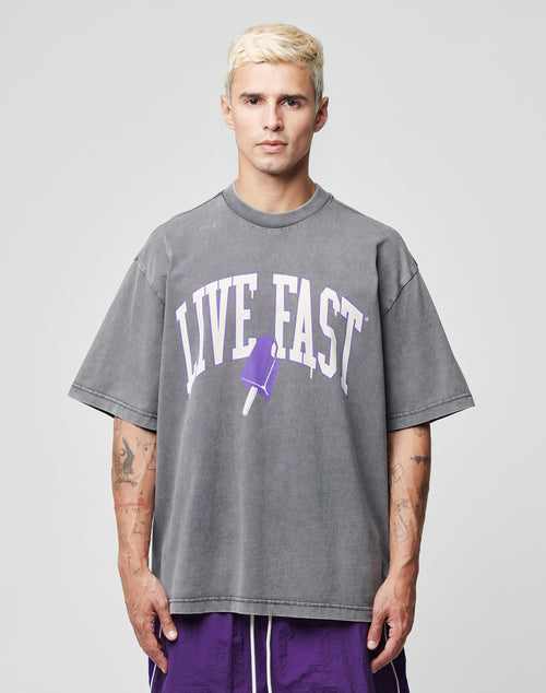 A person with short blond hair wears the LIVE FAST DIE YOUNG Live Fast Ice Tee, an oversized grey 100% Baumwolle shirt with a purple popsicle graphic and LIVE FAST print, paired with purple shorts against a plain light gray background.