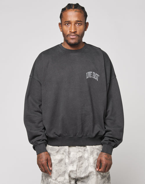 A man with braided hair wears the Live Fast Berlin Sweater by LIVE FAST DIE YOUNG—an oversize black sweatshirt with LIVE FAST text and topstitching, paired with light gray patterned pants. Crafted from 100% Baumwolle for comfort, set against a plain light background.