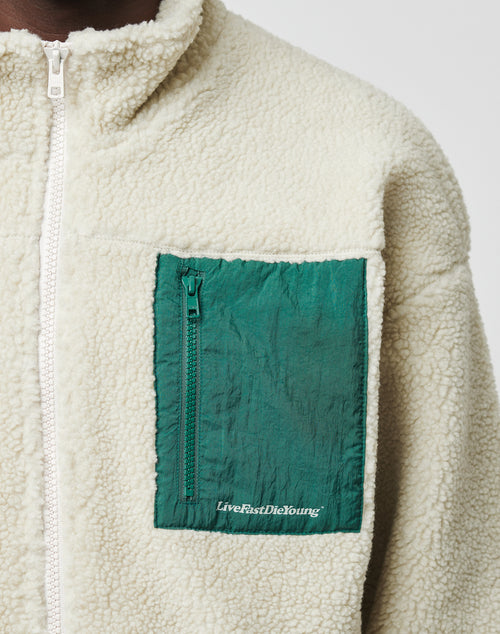 A close-up of the Basic Teddy Jacket by LIVE FAST DIE YOUNG in cream fleece with a standing collar and green zippered chest pocket. LiveFastDieYoung is printed in white near the bottom. Features a white zipper and comfortable regular fit.