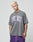 A person with short blond hair wears the LIVE FAST DIE YOUNG Live Fast Ice Tee—an oversize, faded gray 100% Baumwolle shirt with a bold purple and white front print—paired with purple shorts. Arm tattoos are visible against a plain background.