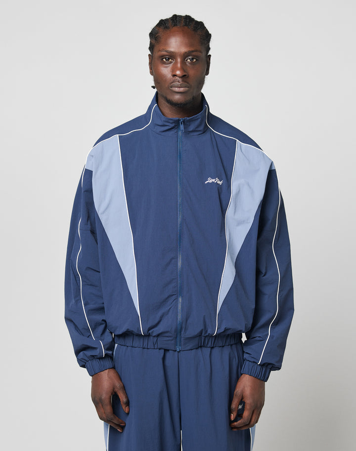 A person stands facing forward in a blue and light blue LIVE FAST DIE YOUNG Basic Nylon Trackjacket with matching pants and elastic cuffs, set against a plain light gray background.