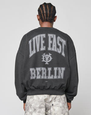 A person with braided hair faces away, wearing the LIVE FAST DIE YOUNG Live Fast Berlin Sweater—an oversized black 100% Baumwolle sweatshirt with bold gray LIVE FAST BERLIN text and graphic, paired with light gray patterned pants.