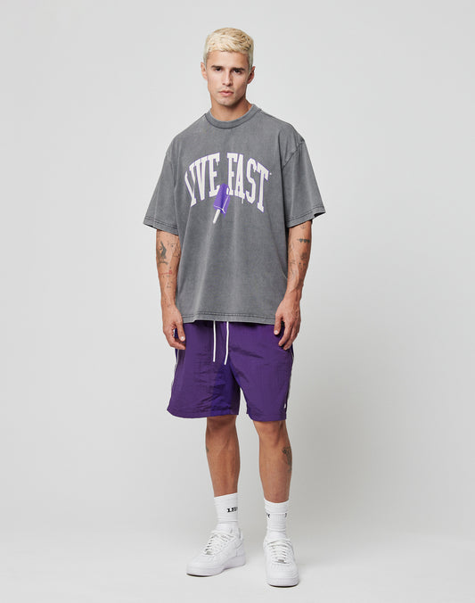A young man with short blond hair wears a gray oversized LIVE FAST DIE YOUNG nylon t-shirt, purple Beach Shorts, white socks, and white sneakers, standing against a plain light gray background.
