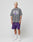 A man with short blond hair faces forward, wearing an oversize grey Live Fast Ice Tee from LIVE FAST DIE YOUNG in 100% cotton, paired with purple shorts, white socks, and white sneakers, posing against a light grey background.