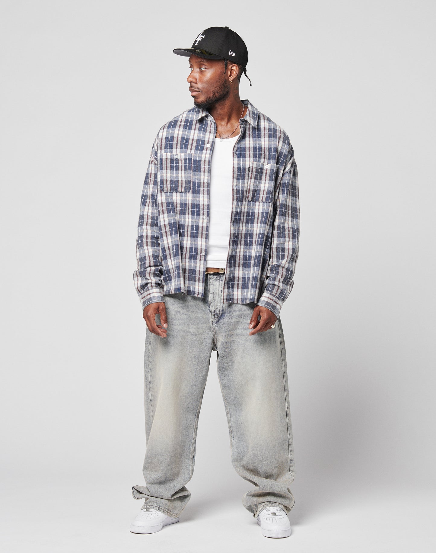 A man in a black cap, blue and white plaid shirt over white tee, LIVE FAST DIE YOUNG LFDY Baggy Denim jeans (baggy fit, faded, wide straight leg), and white sneakers stands slightly turned against a plain gray background.