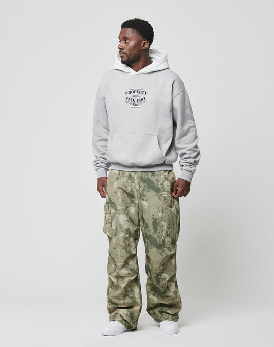A man stands against a plain background wearing the LIVE FAST DIE YOUNG Live Fast Property Hooded sweatshirt, green camouflage cargo pants, and white sneakers. He faces slightly to the side with his hands relaxed by his sides.