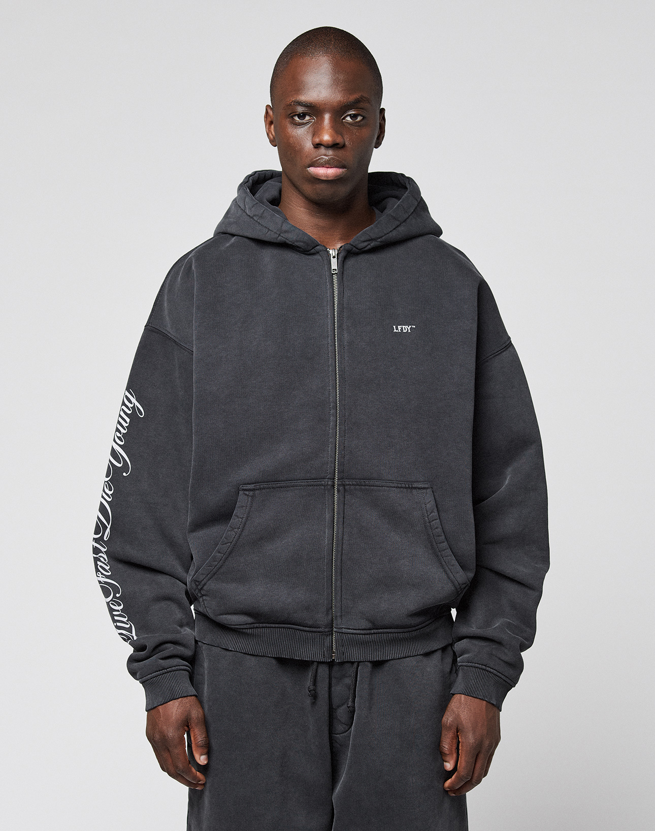 A man wears the LIVE FAST DIE YOUNG LFDY Run Club Hooded Zip and matching sweatpants with Ärmelprint, made from soft Baumwolle Polyester in an oversize fit, featuring small chest lettering, against a plain light gray background.