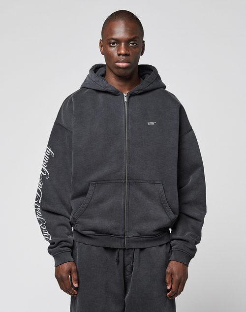 A man wears the LIVE FAST DIE YOUNG LFDY Run Club Hooded Zip and matching sweatpants with Ärmelprint, made from soft Baumwolle Polyester in an oversize fit, featuring small chest lettering, against a plain light gray background.
