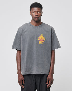A man stands against a plain light gray background wearing the Melting Chest Tee by LIVE FAST DIE YOUNG—a loose-fitting, vintage-wash gray t-shirt featuring a colorful chest graphic.