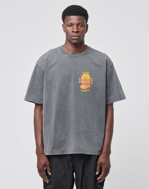 A man stands against a plain light gray background wearing the Melting Chest Tee by LIVE FAST DIE YOUNG—a loose-fitting, vintage-wash gray t-shirt featuring a colorful chest graphic.