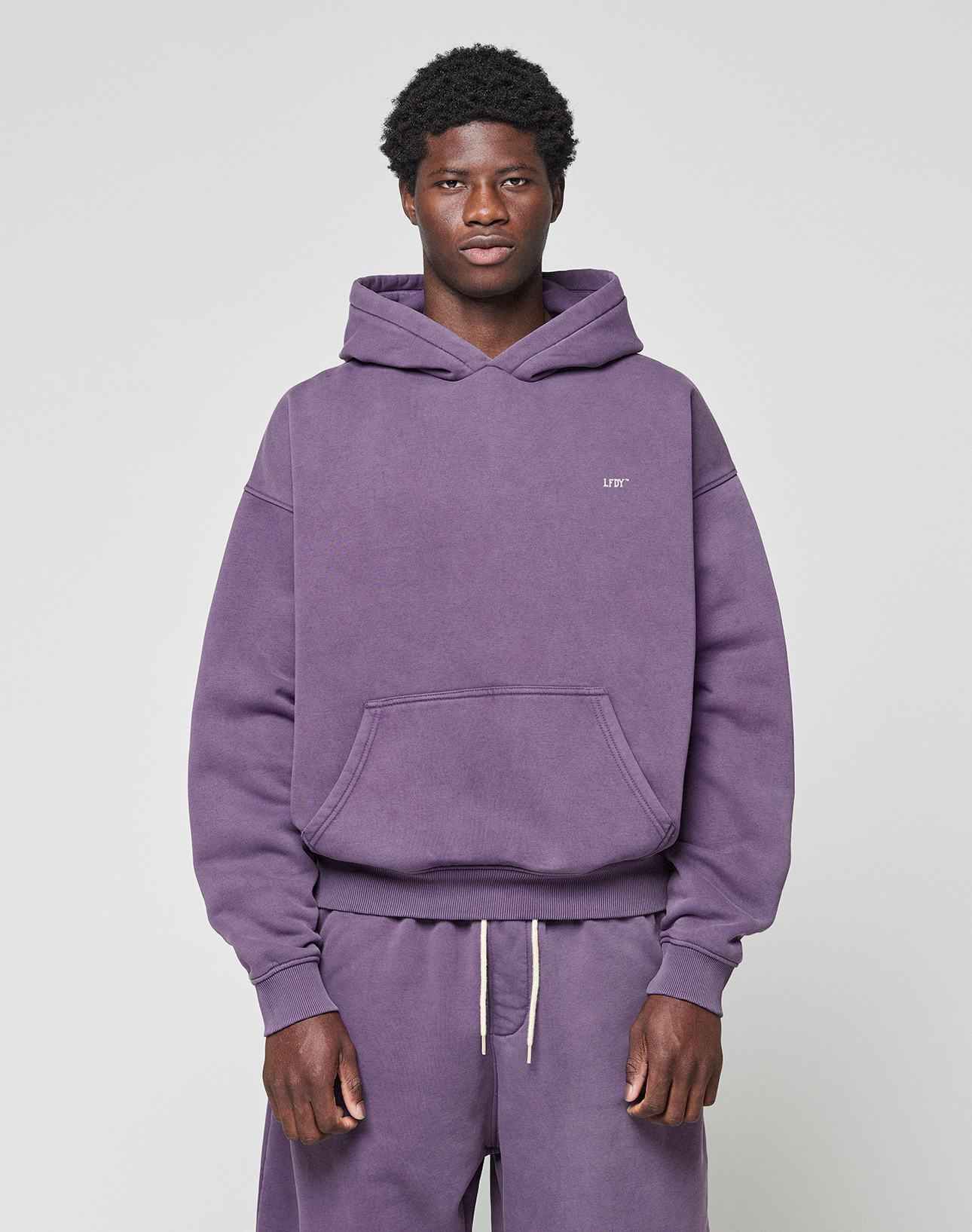 A young man faces forward in a purple LIVE FAST DIE YOUNG Basic Evidence Hooded Oversize Fit hoodie and matching sweatpants. Made from a soft Baumwolle Polyester blend, the hoodie has a small white logo on the left chest. The background is light and plain.