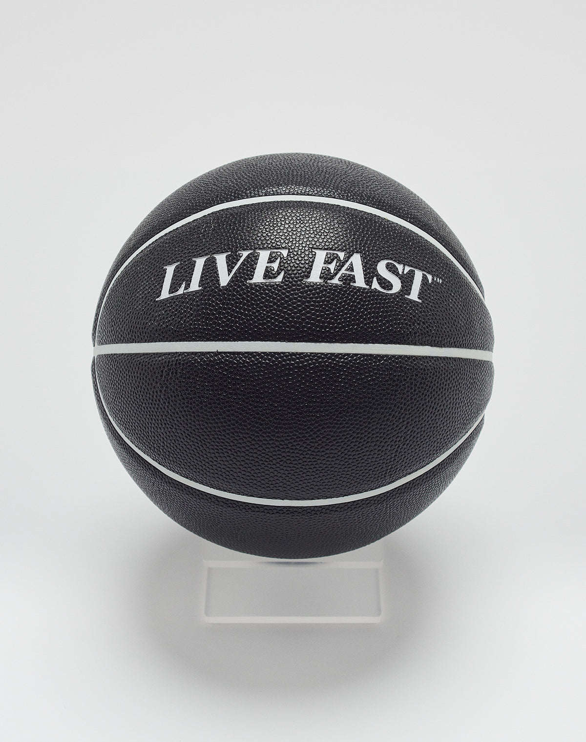 The Live Fast Basketball by LIVE FAST DIE YOUNG features a black design with white stripes, deep grooves, and LIVE FAST branding in hygroscopic PU, displayed on a clear stand against a white background. 