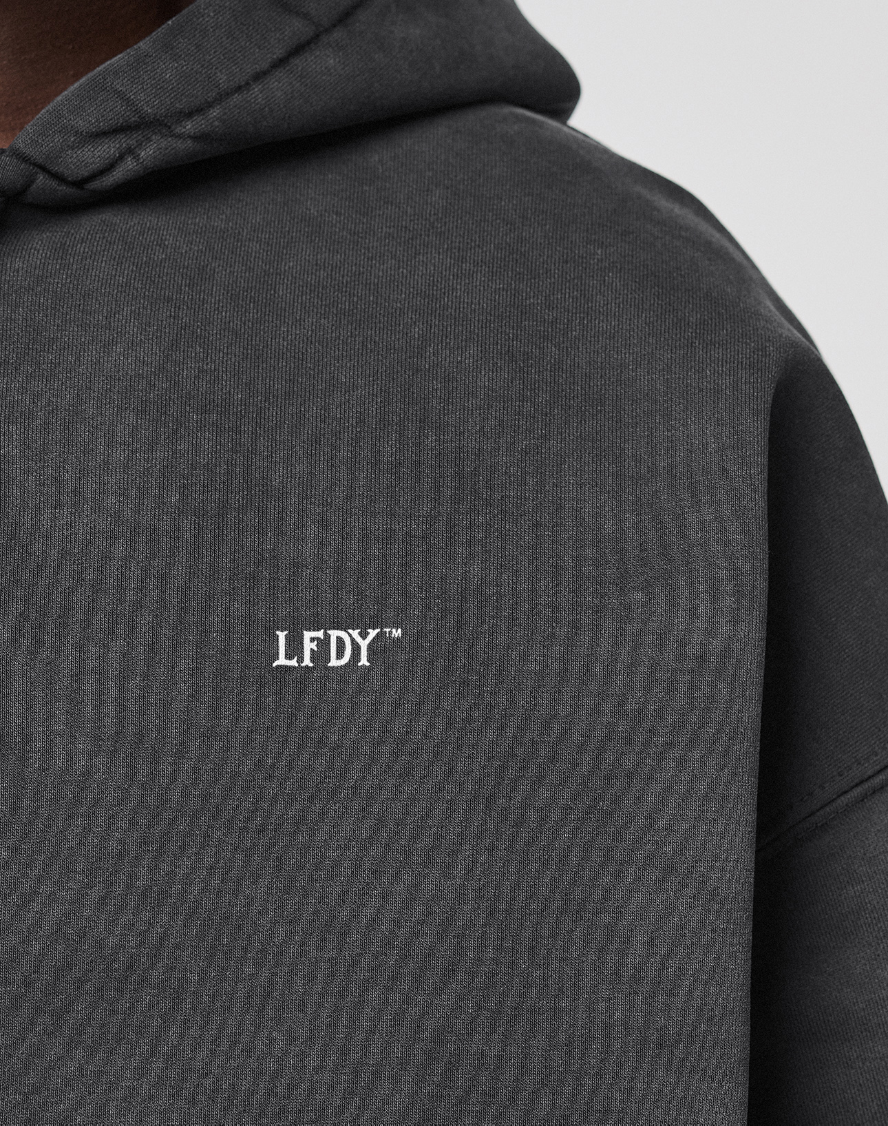 A close-up of the dark gray LIVE FAST DIE YOUNG LFDY Run Club Hooded Zip, featuring white LFDY™ chest embroidery. The oversized fit, made from soft Baumwolle Polyester, partially covers the wearer’s shoulder and neck.