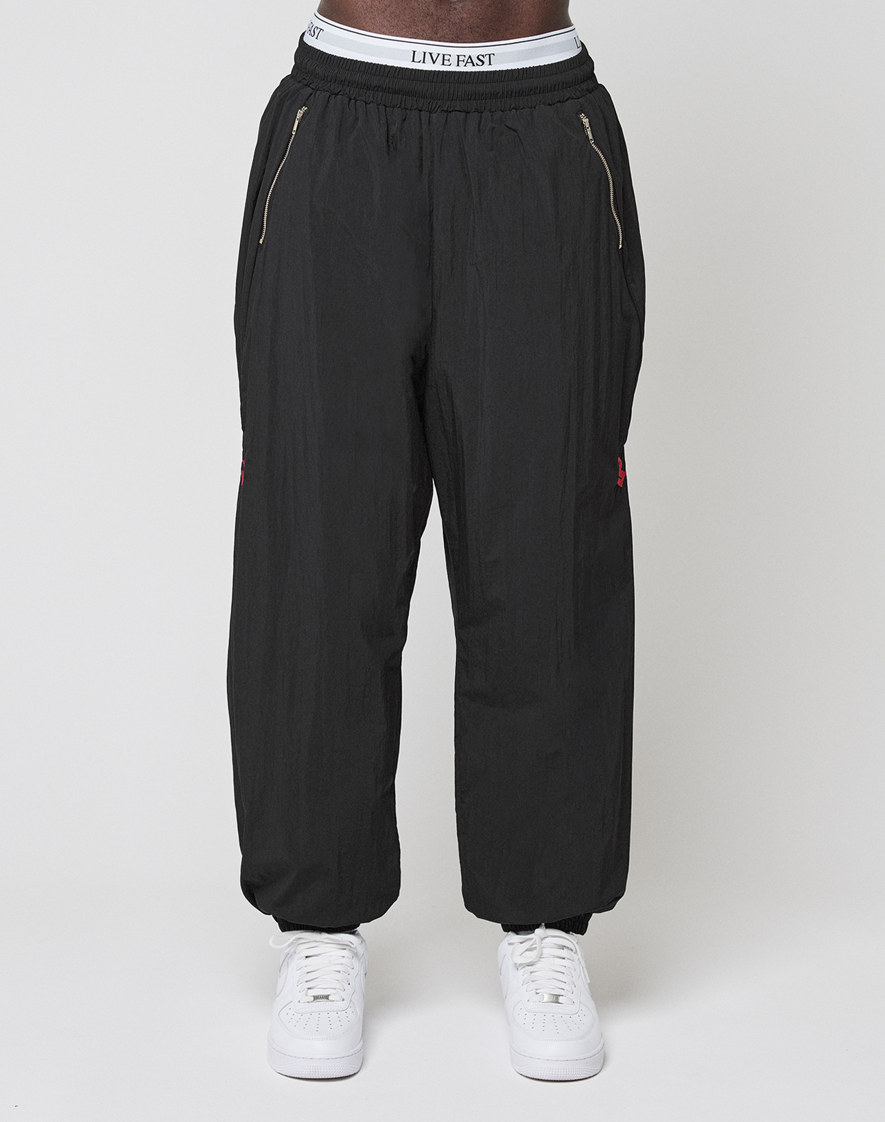 Someone wears the LFDY Dealership Trackpants by LIVE FAST DIE YOUNG, featuring zippered pockets, an elastic waistband with "LIVE FAST" visible, paired with white sneakers on a plain white background.