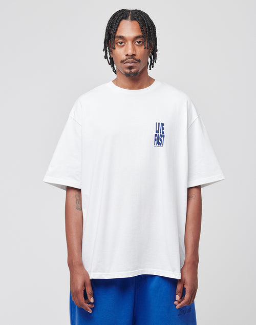 A man with braided hair wears the LIVE FAST DIE YOUNG Live Fast Lenox Tee, an oversized white cotton T-shirt with blue front print, paired with matching blue shorts. He stands facing forward against a plain light gray background.