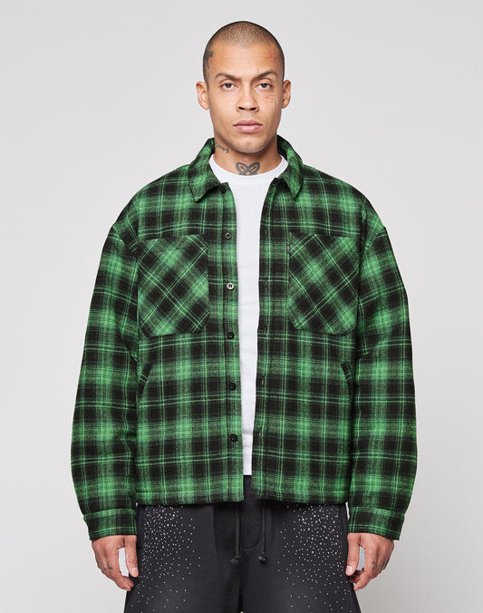A person stands facing forward in a LIVE FAST DIE YOUNG LFDY Padded Flannel—green and black plaid, worn over a white T-shirt with black pants with white speckles. The background is plain and light-colored. | model