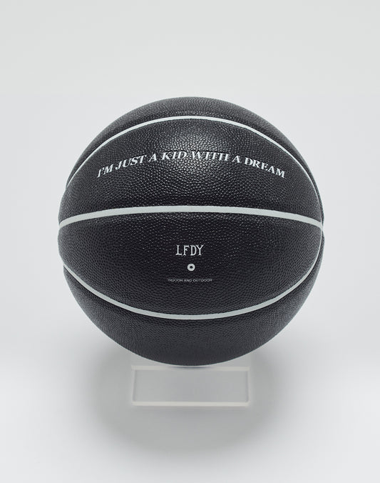 A Live Fast Basketball from LIVE FAST DIE YOUNG, black with white stripes and deep grooves, sits on a clear stand. It features IM JUST A KID WITH A DREAM, the initials LFDY, and small extra text below.