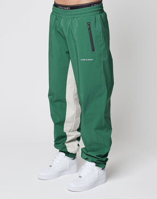 A person wears LIVE FAST DIE YOUNG Basic Shell Pants—green with a vertical white stripe, reflektierender Druck, zippered pocket, oversize fit, Elastikbund mit Kordelzug and LIVE FAST text on the waistband—plus white sneakers. Background is plain white.