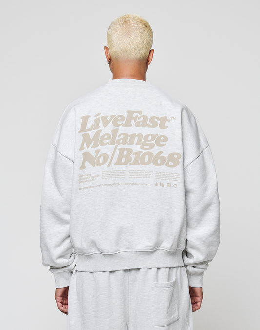 A person with short platinum blonde hair wears the LIVE FAST DIE YOUNG Basic Melange Sweater, an oversized light gray cotton sweatshirt featuring large beige Live Fast text on the back, standing against a plain light background.