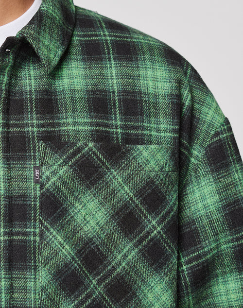 Close-up of a person wearing the LIVE FAST DIE YOUNG LFDY Padded Flannel, an oversized green and black plaid overshirt with chest pockets, a small tag on the pocket seam, and a white undershirt visible for a relaxed style.