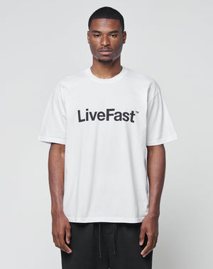A man faces forward, wearing the Basic LiveFast Tee by LIVE FAST DIE YOUNG, a loose white T-shirt with LiveFast in bold black letters on the chest. The Regular Fit shirt is photographed against a plain, light background.