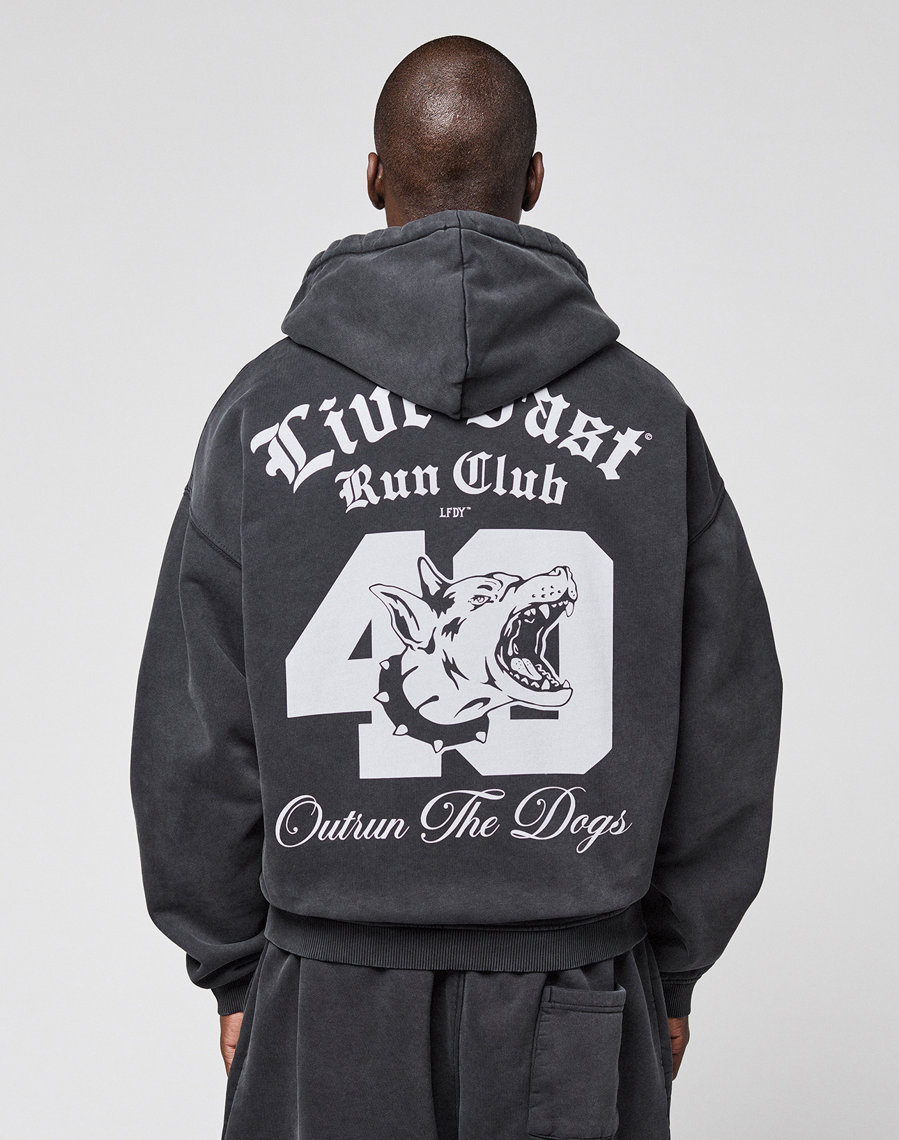 Someone wearing the LIVE FAST DIE YOUNG LFDY Run Club Hooded Zip, a black oversized hoodie featuring bold white graphics on the back: "Live Fast Run Club," "49," "Outrun The Dogs," and a snarling dog head. Made from cotton-polyester.