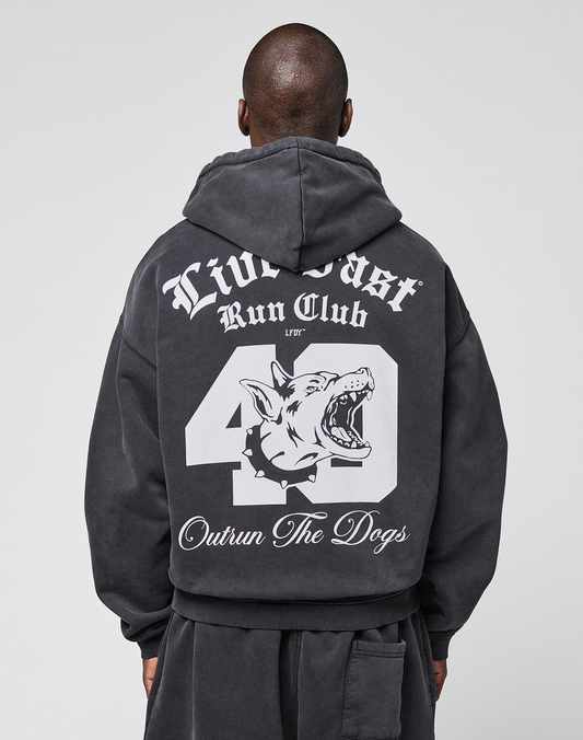 Someone wearing the LIVE FAST DIE YOUNG LFDY Run Club Hooded Zip, a black oversized hoodie featuring bold white graphics on the back: "Live Fast Run Club," "49," "Outrun The Dogs," and a snarling dog head. Made from cotton-polyester.