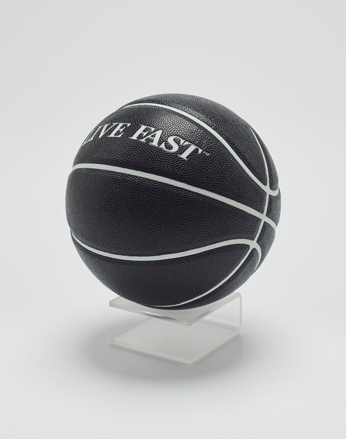 The Live Fast Basketball by LIVE FAST DIE YOUNG is black with deep grooves and white lines, featuring LIVE FAST branding. It is displayed on a clear stand against a plain white background.