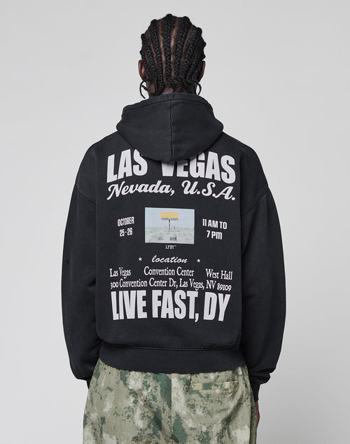 A person with braided hair wears the LIVE FAST DIE YOUNG Live Fast Vegas Hooded with white front and back print plus a small photo for an event at the Las Vegas Convention Center, paired with green camouflage pants.