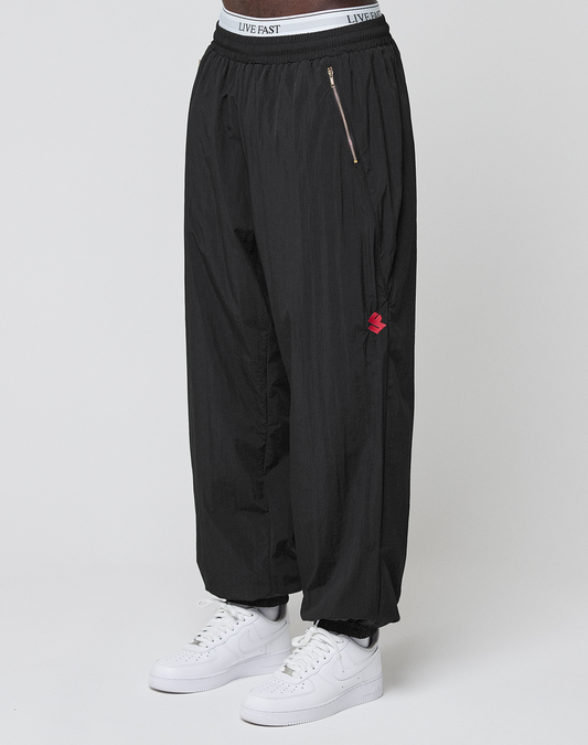 A person stands against a plain background wearing LFDY Dealership Trackpants by LIVE FAST DIE YOUNG—loose black track pants with an elastic waistband labeled LIVE FAST and a zipper pocket—paired with white Nike sneakers.