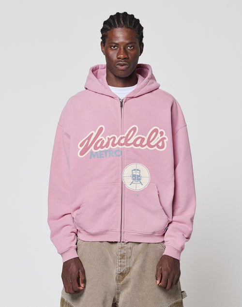 A man faces the camera in a neutral pose, wearing the LIVE FAST DIE YOUNG LFDY Vandals Hooded Zip in light pink with a “Vandals Metro” front print, paired with beige pants, against a plain background.