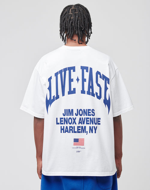 A person with braided hair is wearing the LIVE FAST DIE YOUNG Live Fast Lenox Tee, an oversized white cotton T-shirt with a bold LIVE FAST front print and Jim Jones Lenox Avenue Harlem, NY plus a small American flag on the back.