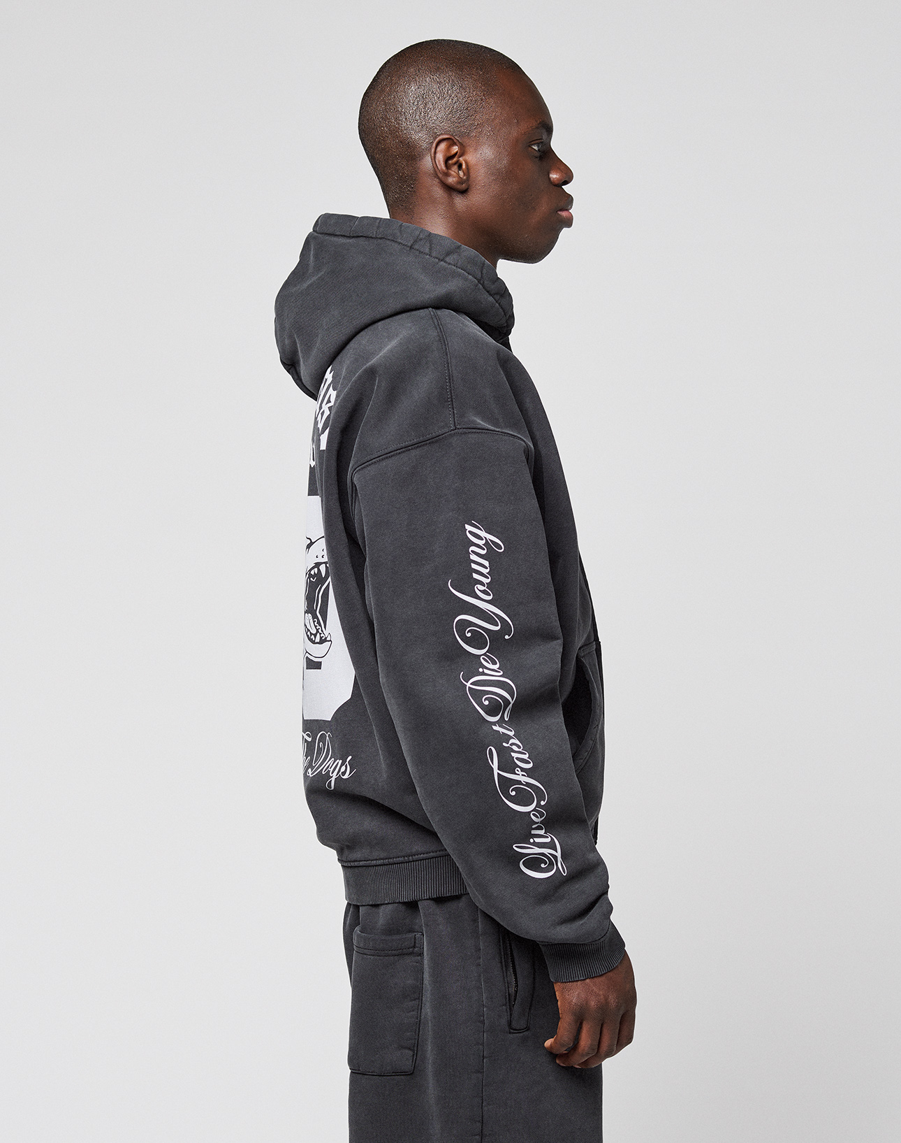 A person stands in profile wearing the LIVE FAST DIE YOUNG LFDY Run Club Hooded Zip and matching pants. The hoodie features a white sleeve print, a large back graphic, and is made from a soft cotton-polyester blend. Plain light background.