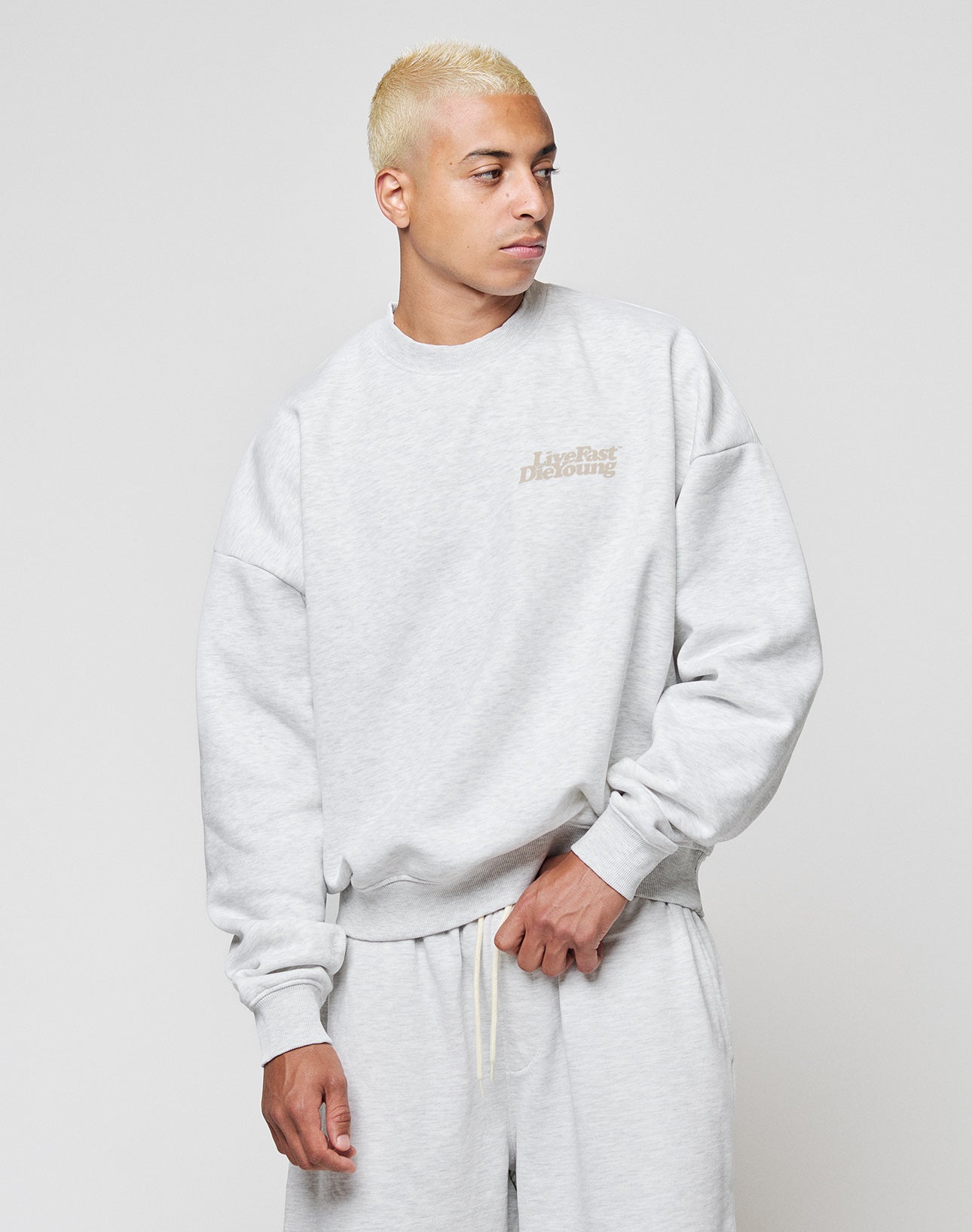 A person with short, bleached blonde hair wears the LIVE FAST DIE YOUNG Basic Melange Sweater and matching pants in a relaxed fit, made from a soft cotton-poly blend. They look to the side, one hand on the hem, against a plain light background.