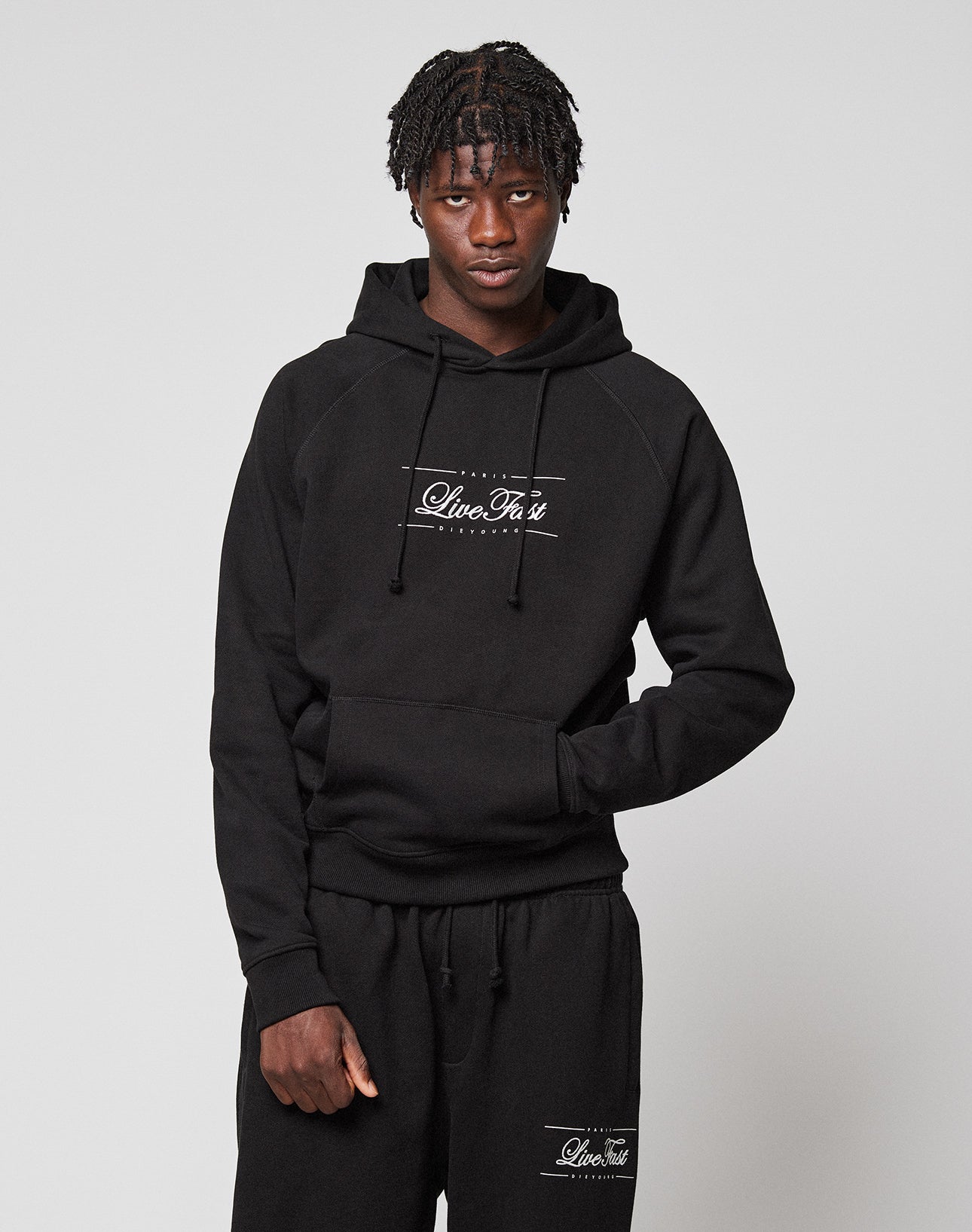 A young man with short braids models the LIVE FAST DIE YOUNG LFDY Embroidery Hooded and matching black sweatpants with white LiveVäst text, made from soft French terry, standing against a plain light gray background.