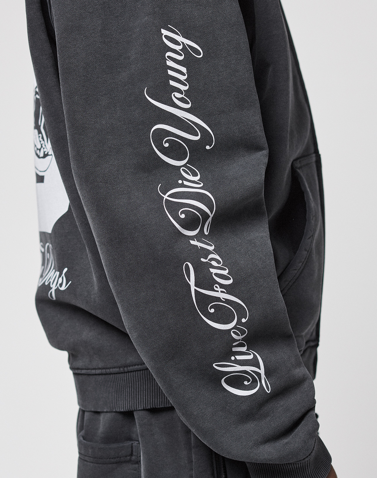 Wearing the LFDY Run Club Hooded Zip by LIVE FAST DIE YOUNG, a person poses with their hand in their pocket. The black oversize sweatshirt features a white cursive Ärmelprint reading "Live Fast Die Young." Only part of their torso and arm is visible.