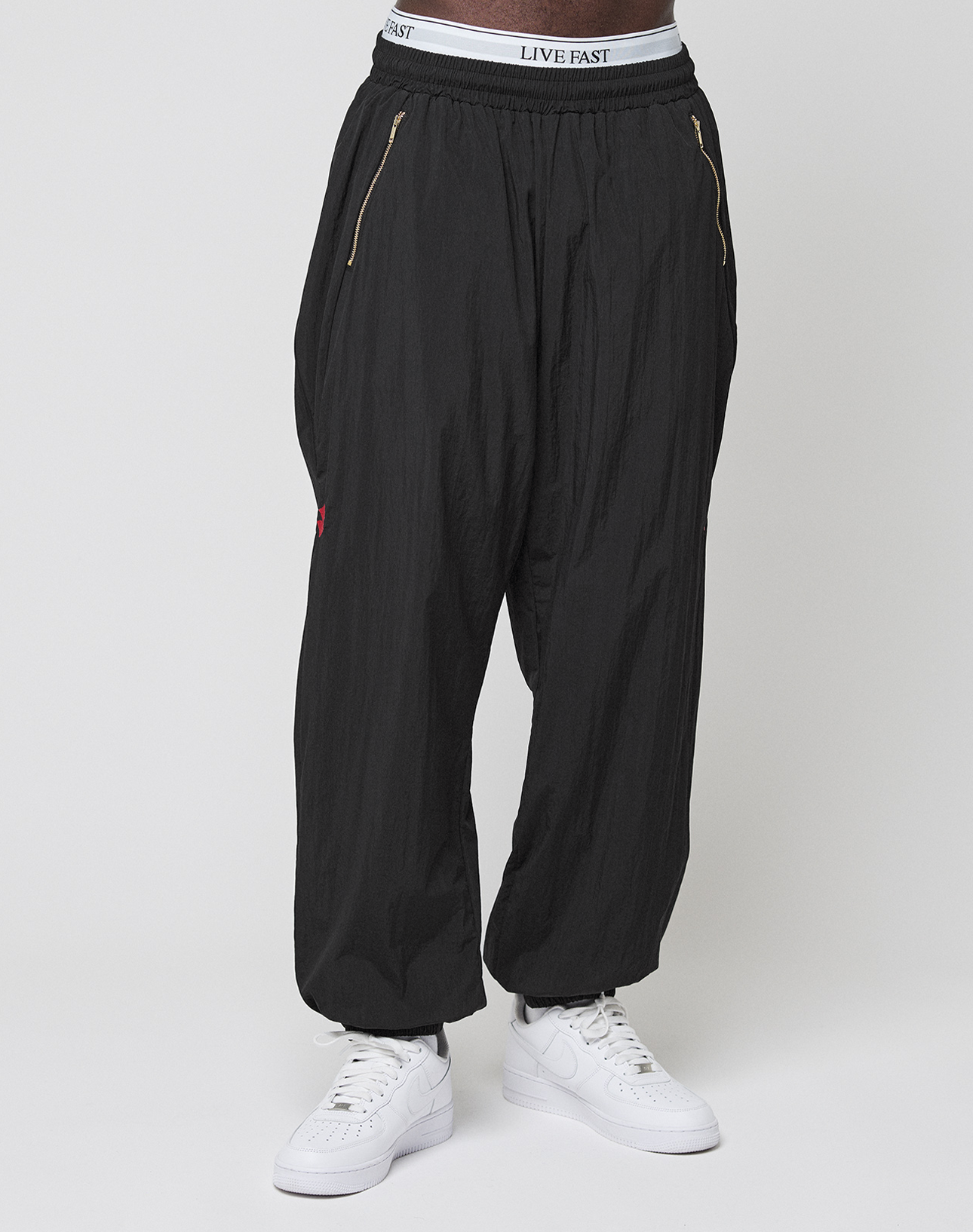 A person models the LIVE FAST DIE YOUNG LFDY Dealership Trackpants—loose-fitting black pants with gold zippers, a white LIVE FAST waistband, and white sneakers. The wide fit, elastikbund, and polyamide fabric ensure sporty comfort.
