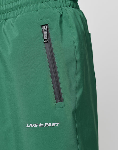 Close-up of Basic Shell Pants by LIVE FAST DIE YOUNG: green athletic pants with elastic waistband and drawstring, a black zippered pocket, and LIVE FAST in reflective white print near the pocket.