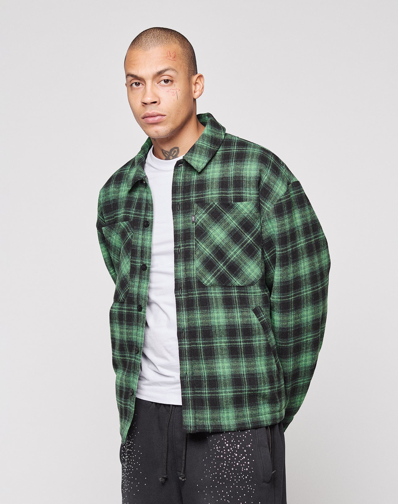 A person with a shaved head wears the green and black LIVE FAST DIE YOUNG LFDY Padded Flannel over a white t-shirt and black pants, standing against a plain light gray background.