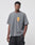 A man with short hair and a neutral expression wears the LIVE FAST DIE YOUNG Melting Chest Tee, an oversize faded gray cotton T-shirt with a small yellow and red chest graphic, paired with black cargo pants against a plain background.