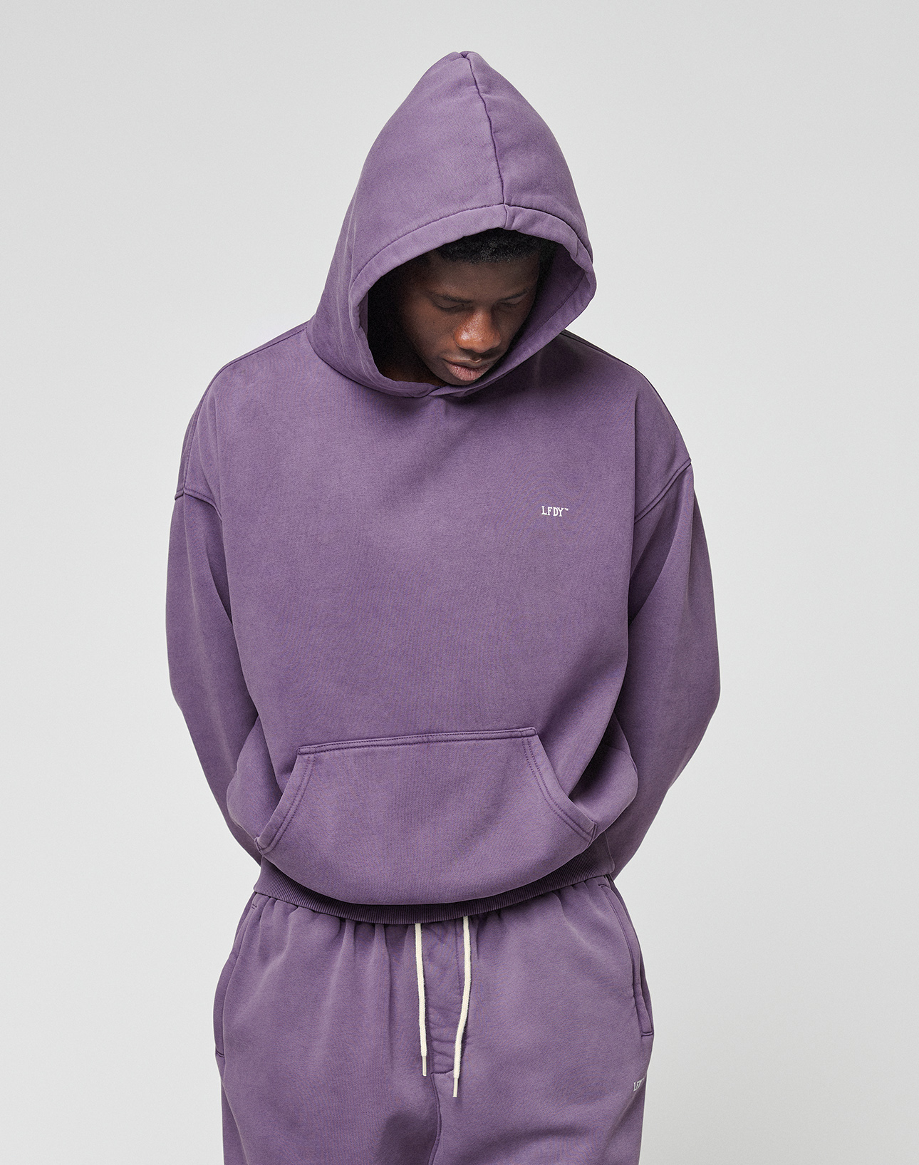 A person wearing the LIVE FAST DIE YOUNG Basic Evidence Hooded oversize purple cotton-polyester sweatshirt and matching sweatpants stands against a plain light background, hood up, looking down with hands behind their back.