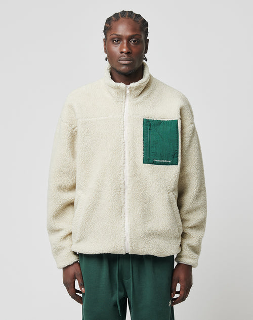 A man faces forward wearing the LIVE FAST DIE YOUNG Basic Teddy Jacket in cream with a Stehkragen and green nylon zippered breast pocket, paired with dark green pants against a plain light gray background.