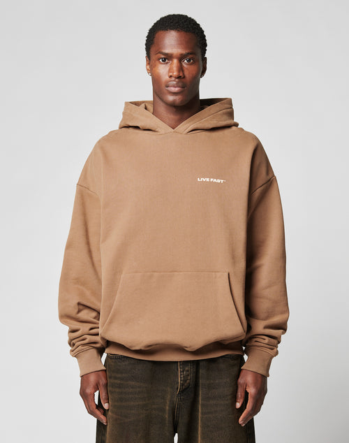 A man stands facing forward, hands by his sides, wearing a brown Live Fast Plain Hooded sweatshirt from LIVE FAST DIE YOUNG, featuring an oversize fit and small white LIVE FAST text on the chest against a plain light background.