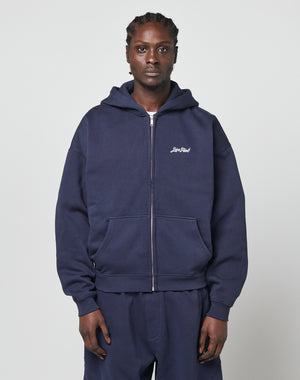 A person wears a navy blue LIVE FAST DIE YOUNG Basic Script Hooded Zip and matching sweatpants, facing forward against a plain light background. The soft Baumwolle hoodie features small white text on the left chest.