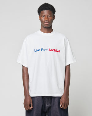 A man stands against a plain light gray background wearing the LIVE FAST DIE YOUNG Live Fast Archive Tee, an oversize white T-shirt featuring Live Fast Archive printed in blue and red on the front.