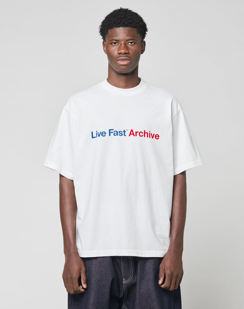 A man stands against a plain light gray background wearing the LIVE FAST DIE YOUNG Live Fast Archive Tee, an oversize white T-shirt featuring Live Fast Archive printed in blue and red on the front.