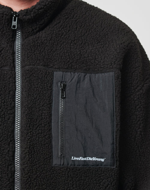 Close-up of the LIVE FAST DIE YOUNG Basic Teddy Jacket in black fleece with a textured surface, Regular Fit, and a black nylon chest pocket featuring “LiveFastDieYoung” printed in white.