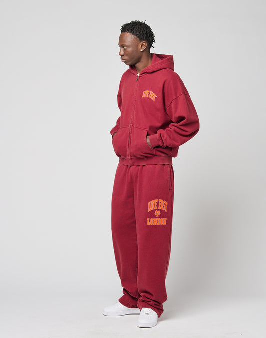 A man stands against a plain background wearing the LIVE FAST DIE YOUNG “Live Fast London Hooded Zip” and matching maroon sweatpants with yellow front and back prints, made from 100% cotton, hands in pockets, styled with white sneakers.