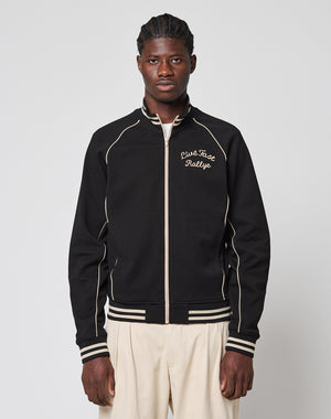 A man stands facing forward, wearing the Live Fast Tracksuit Jacket—a black zip-up with white piping, Eingrifftaschen, and a Piste Face Rallye Chainstitch Logo on the chest—paired with light pants against a plain light background.